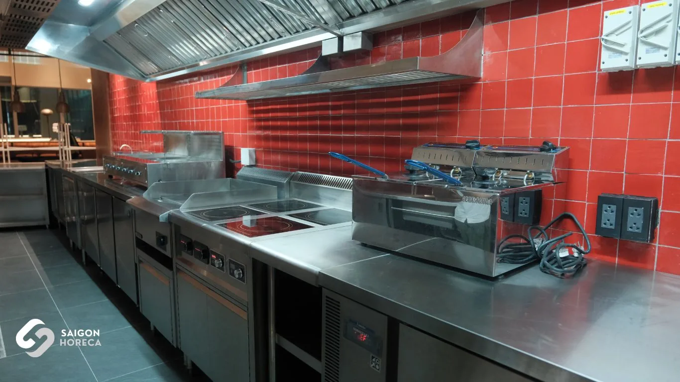 Commercial stainless steel cooking range at Mam Mam: induction stoves, gas ranges, and exhaust canopy hood set against the red brick backdrop