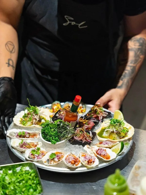 SOL service staff presenting freshly shucked oysters on the half shell with shallots, fresh herbs, and salsa, accompanied by lime wedges and hot sauce in signature Latin American fashion