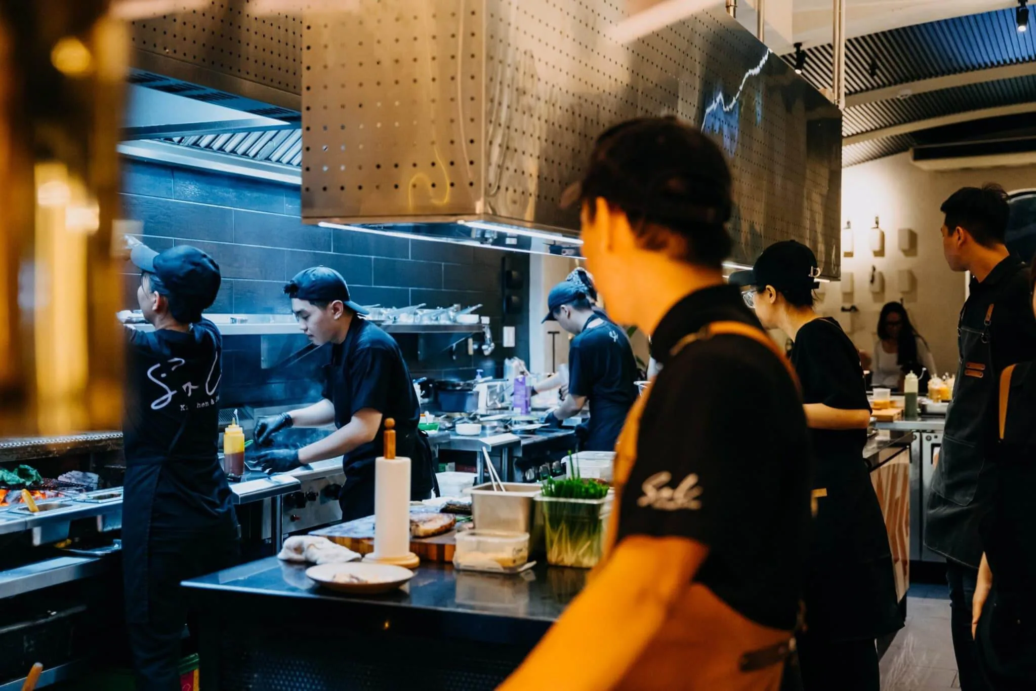 The open theater kitchen of Sol Kitchen & Bar featuring chefs in black uniforms and orange aprons cooking on heavy-duty stainless steel cooking lines beneath an industrial exhaust hood