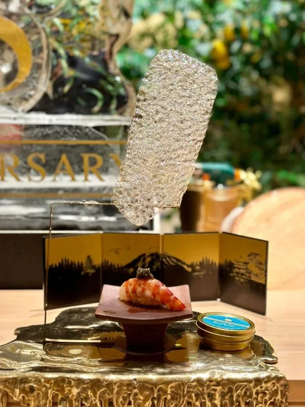 Amaebi sweet shrimp nigiri garnished with edible gold leaf and caviar before a vintage Japanese landscape folding screen